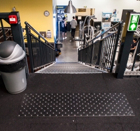 Vue du haut d’un escalier ERP dans une salle de sport avec revêtement de sécurité et indication de sortie de secours
			